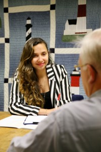 A woman with long curly hair wearing a black and white striped blazer is engaged in a conversation with an older man.