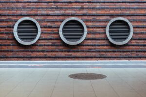 Three circular vents on a red brick wall above a tiled floor with a round metal grate.